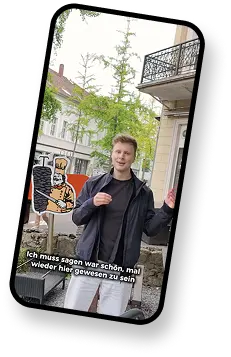 Ein junger Mann steht draußen vor einem Gebäude und lächelt. Links von ihm ist ein Cartoon-Döner abgebildet. Im unteren Bereich des Bildes ist ein Text überlagert.