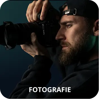 Ein bärtiger Mann mit einer Basecap hält eine professionelle Kamera und schaut ernst durch den Sucher. Das Bild wird von dem fetten Text FOTOGRAFIE überlagert.