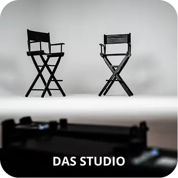 Zwei schwarze Regiestühle stehen auf einem weißen Hintergrund in einem Fotostudio. Im Vordergrund, verschwommen, ist ein weiterer Bereich des Studios zu sehen, mit dem Text „DAS STUDIO“.